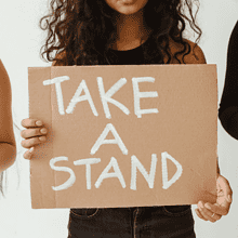 Three individuals are holding protest signs. One sign, held by a person in the center, reads "TAKE A STAND". The individuals are dressed in black, and the plain white background emphasizes the seriousness of their cause. Only their torsos and hands are visible, making their stance on abortion clear yet powerful.