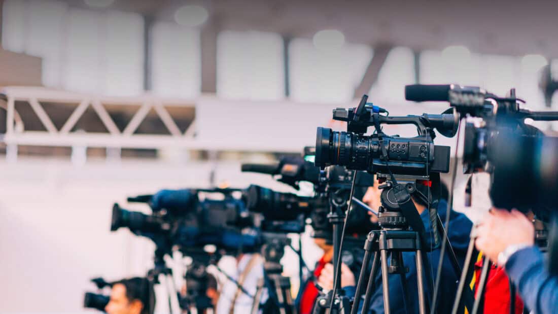 Several cameras are set up on tripods, lined up side by side, focusing on an event. The well-lit indoor area with a blurred background suggests a professional media or press gathering, likely aimed at providing equitable coverage of a significant event like the Olympics.