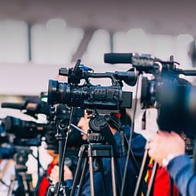 Several cameras are set up on tripods, lined up side by side, focusing on an event. The well-lit indoor area with a blurred background suggests a professional media or press gathering, likely aimed at providing equitable coverage of a significant event like the Olympics.