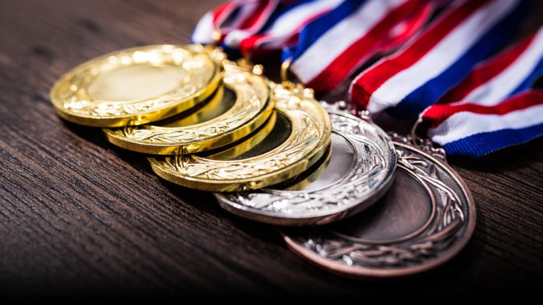 A close-up of several gold, silver, and bronze medals with red, white, and blue ribbons laid out on a wooden surface. These medals have intricate designs etched around the edges.